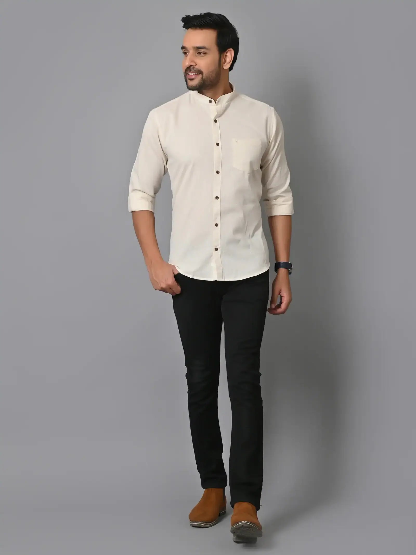Stylish man in cream mandarin-collar shirt, black slim-fit pants, and brown casual shoes posing against a plain gray backdrop, showcasing modern fashion and casual elegance.