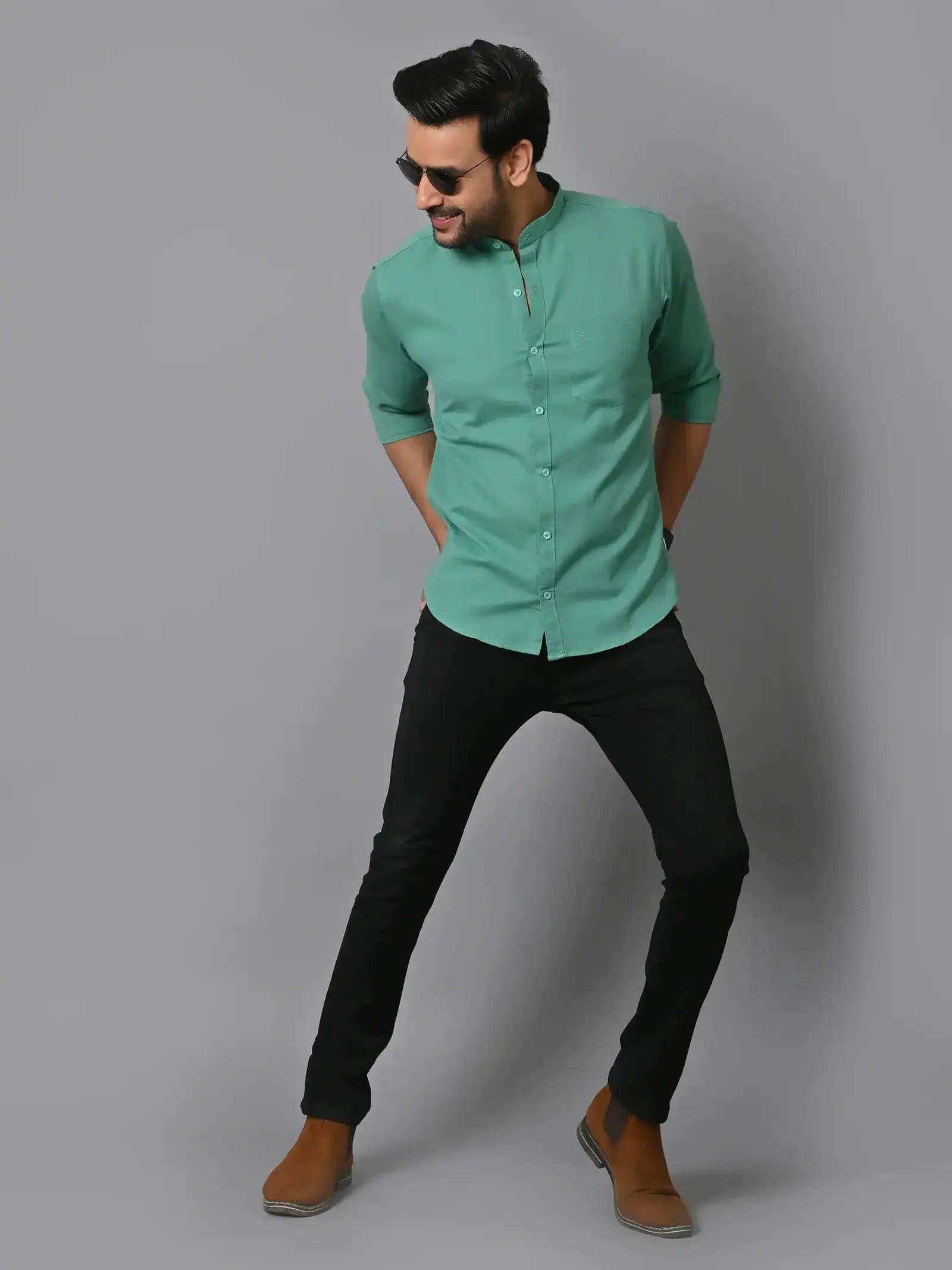 Men's casual fashion model in sunglasses wearing green shirt and black jeans standing against gray background, trendy and stylish menswear photography, CRAFTEARTH.