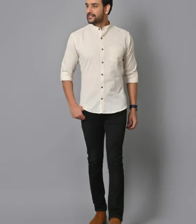 Stylish man in cream mandarin-collar shirt, black slim-fit pants, and brown casual shoes posing against a plain gray backdrop, showcasing modern fashion and casual elegance.